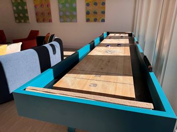 A shuffleboard table with blue and black trim is in the foreground of a room with colorful polka dot wall art.